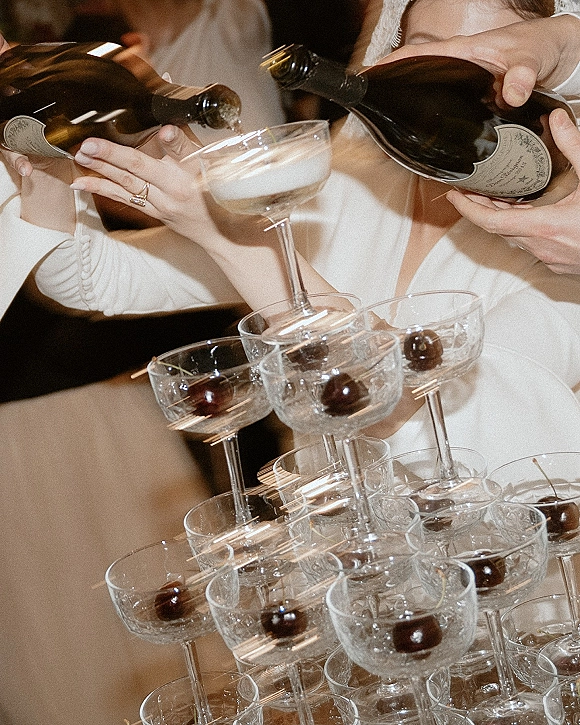 Champagne tower at a wedding as bridal hands pour from a bottle into stacked coupe glasses, cherry garnish and rings in view under warm reception lighting