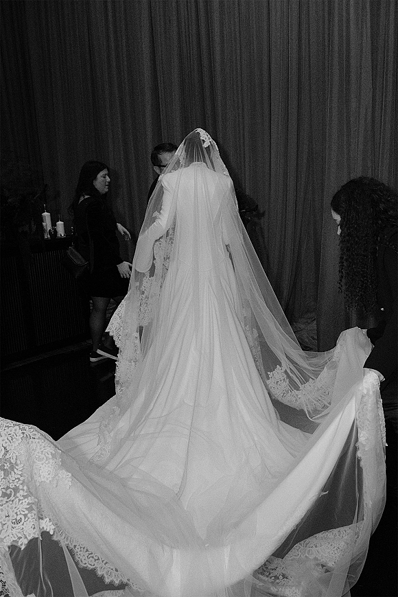 Bridal gown train with cathedral veil and lace trim flowing behind the bride, set against a dark curtain backdrop indoors
