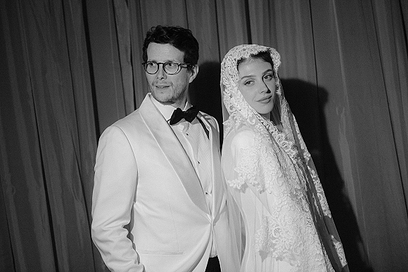 Couple portrait in a black and white wedding portrait style, bride in lace veil and gown with groom in tuxedo and glasses before draped curtains