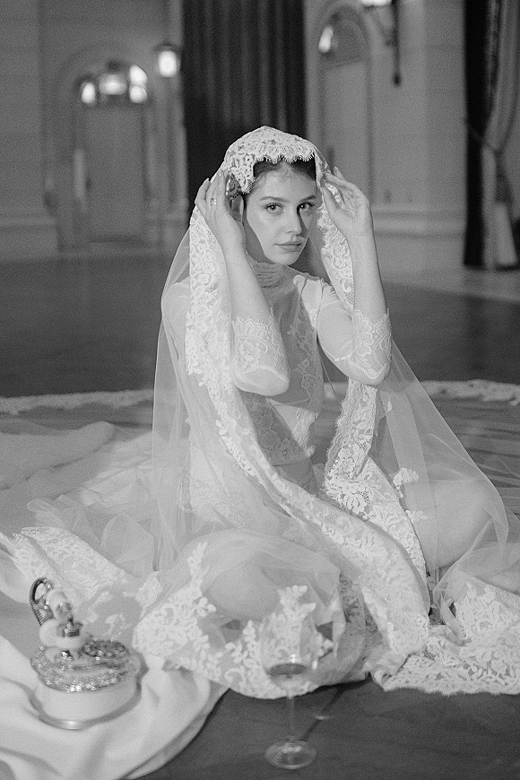 Bridal portrait of a bride in lace veil, seated on a stone floor with long-sleeve lace dress and tiara in an elegant indoor hall.