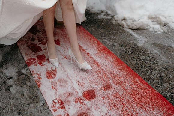 Bridal shoes in a wedding heels close-up, white pointed-toe pumps with lace details peeking beneath the dress hem on a red carpet in snow