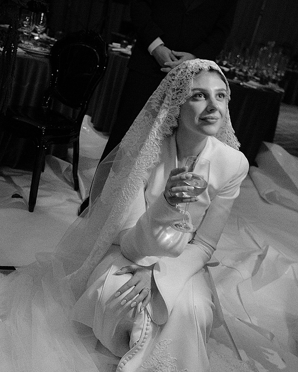 Bridal portrait, black and white wedding portrait of a bride in a white suit with lace veil, holding a wine glass by a reception table