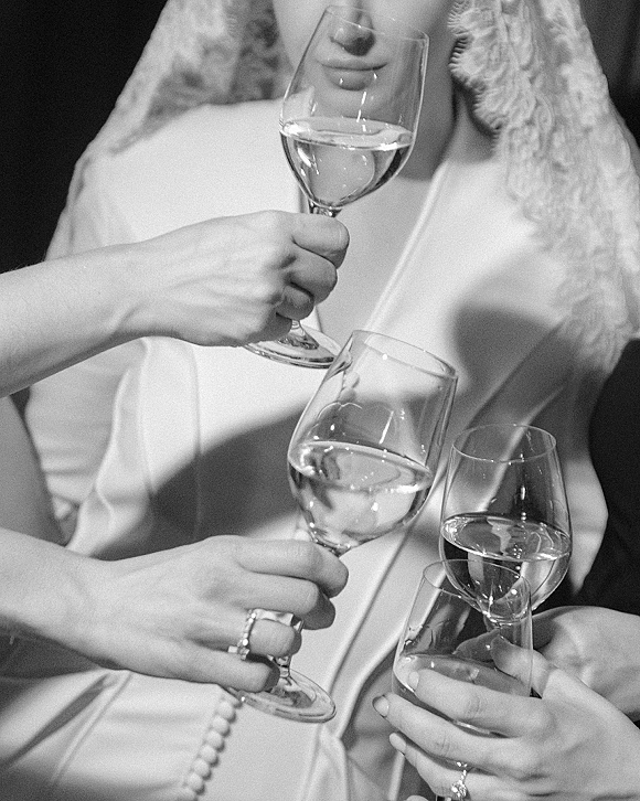 Wedding toast with clinking wine glasses of white wine, showing bride’s lace veil and wedding ring against a dark indoor background