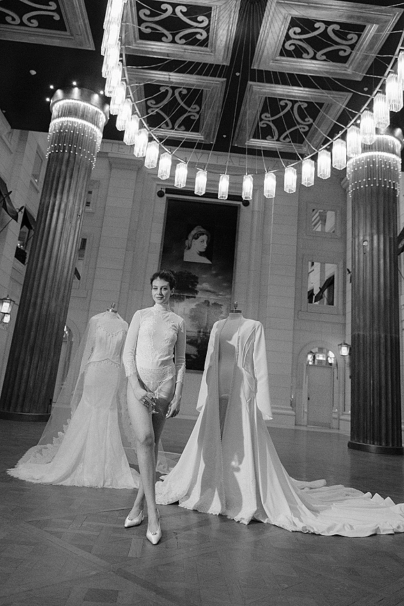 Bridal fashion portrait in a wedding dress showroom, model in a lace bodysuit and heels holding a champagne coupe in a chandelier ballroom
