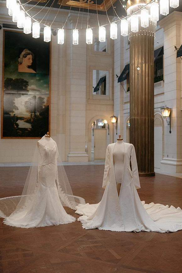 Wedding dresses on mannequins with a cathedral veil and lace long sleeves, displayed in a grand atrium with pendant lights and columns