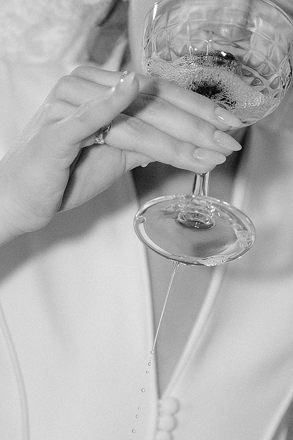 Wedding toast as bride raises a champagne coupe, engagement ring and manicured nails in focus against her wedding dress torso backdrop