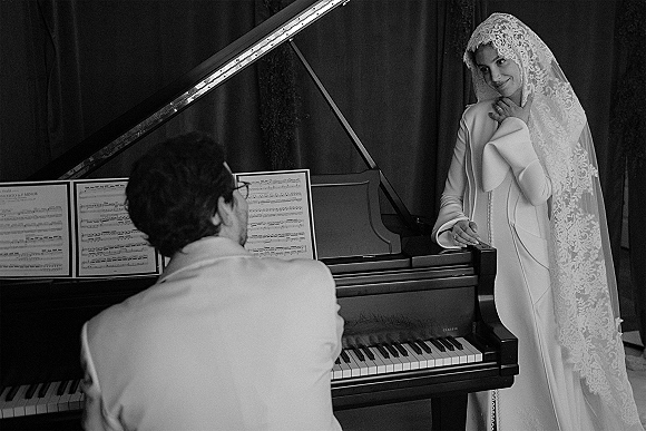 Bride portrait in a lace veil, leaning on a grand piano with sheet music, wearing a long-sleeve gown and ring in a curtained room