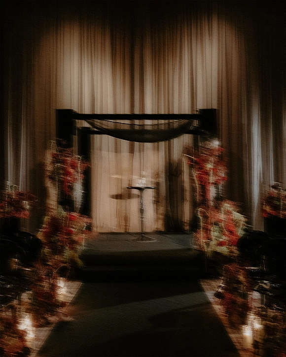 Ceremony altar decor with a black wedding arch, draped fabric, and floral arrangements, set before sheer curtains with candlelit aisle florals indoors