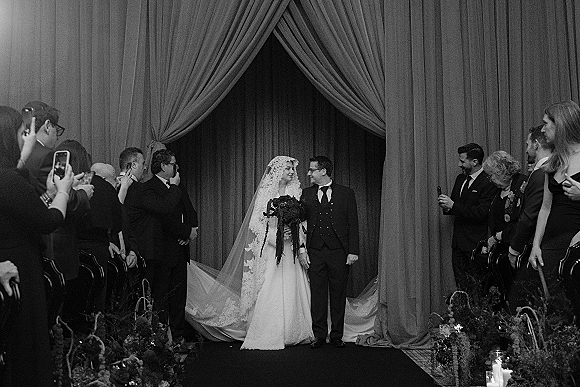 Recessional moment as bride and groom walk the aisle holding hands, her lace veil trailing past candles and guests filming by draped curtains