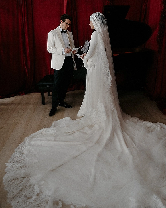 Wedding vows as groom in tuxedo reads from vow papers to bride in lace veil and long train beside a grand piano, red curtains behind