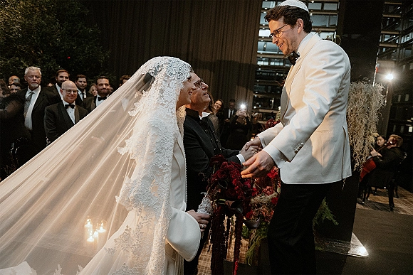 Wedding ceremony moment as bride in lace mantilla veil and long train holds groom’s hand beside deep red florals and candlelit aisle indoors