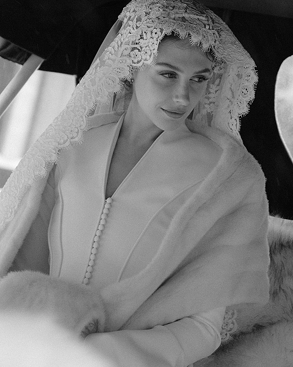 Bridal portrait in black and white of a bride in a lace veil, wearing a shawl and buttoned gown, lit by window light in a car interior