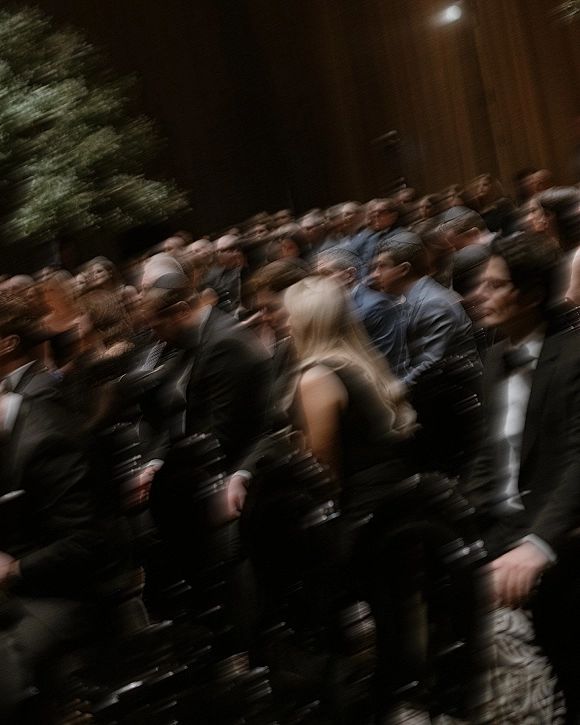 Wedding ceremony guests in formalwear seated in rows of ceremony chairs, watching indoors against a wood-paneled wall with greenery