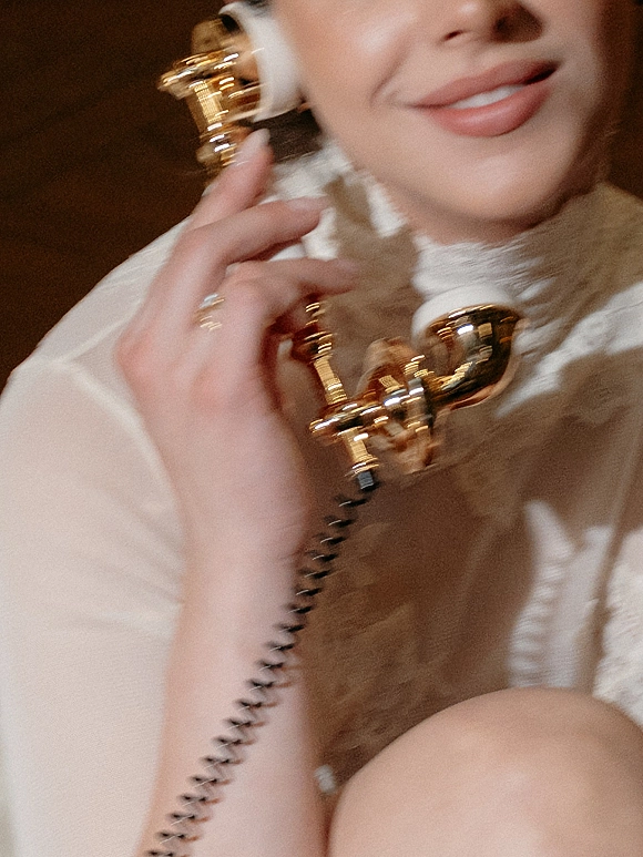 Bridal portrait of a bride holding a vintage phone, showing white lace high-neck top, wedding ring, and nude lipstick against a dark interior background