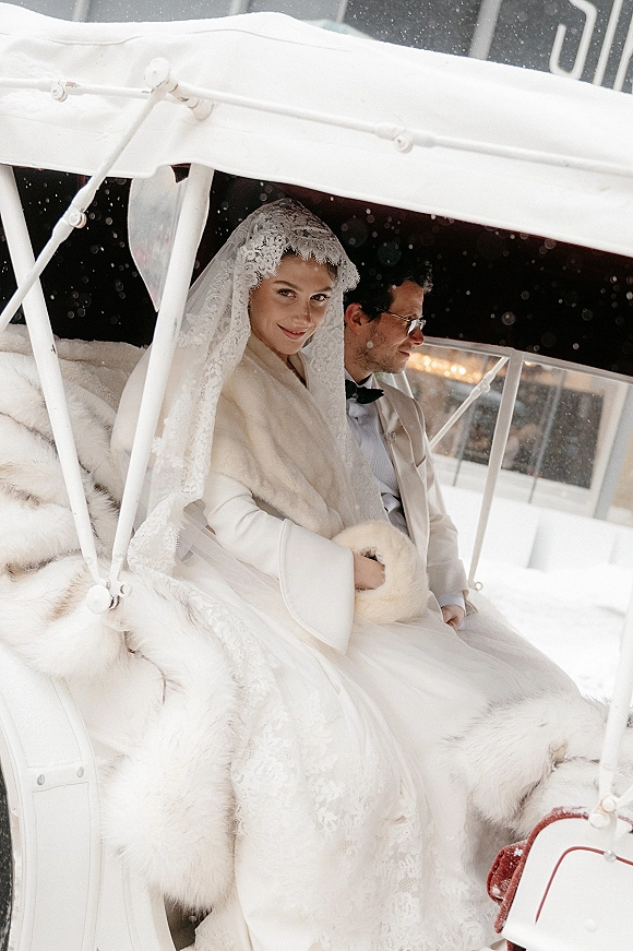 Couple portrait of bride in lace veil and fur stole with groom in tuxedo and glasses, sitting in a carriage in the snow