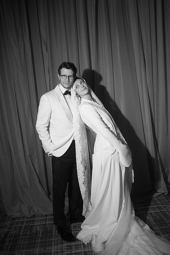 Couple portrait, black and white wedding portrait of bride in lace-trim veil and cape gown leaning beside groom in white tux with glasses by curtains