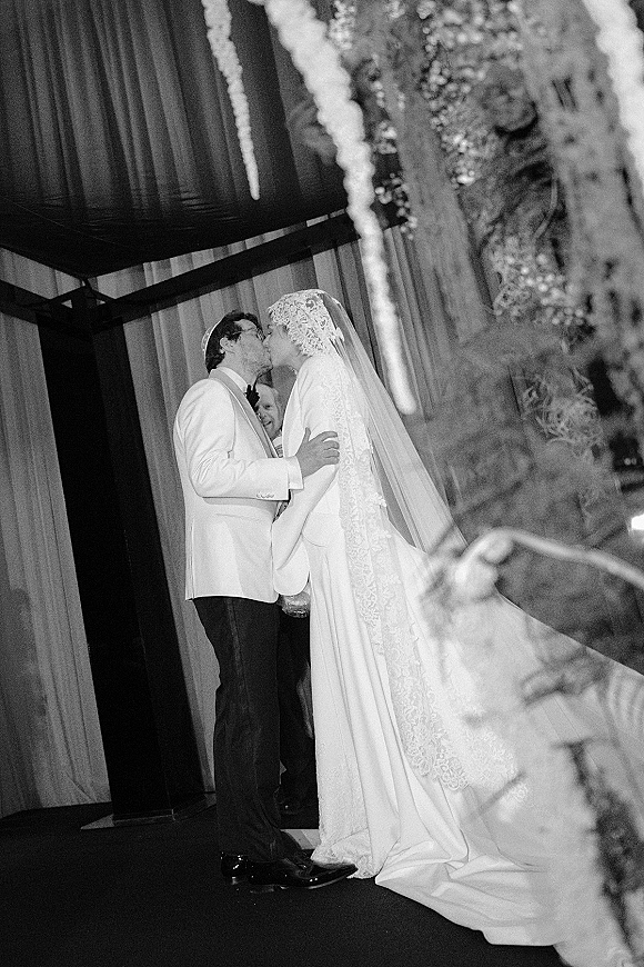 Wedding kiss as bride and groom kissing on an indoor stage, her lace veil and long-sleeve dress train framed by draped curtains and floral garlands