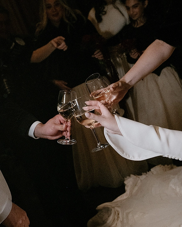 Wedding toast with champagne glasses clinking, hands showing wedding rings and suit sleeve beside a white blazer in dim reception light
