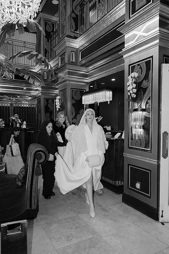 Bridal entrance as bride walks indoors in a wedding dress with cape and long veil, passing a hotel lobby with chandelier and bar counter