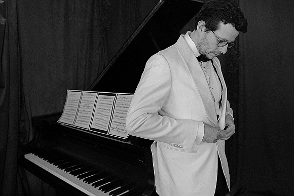 Groom portrait in a white tuxedo jacket and black bow tie, wearing glasses beside a grand piano with sheet music and dark curtain backdrop