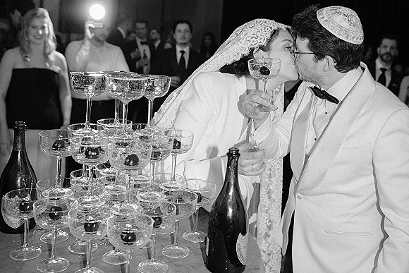 Wedding kiss as newlyweds kissing beside a champagne tower of coupe glasses, bride in veil and lace headpiece, guests behind indoors