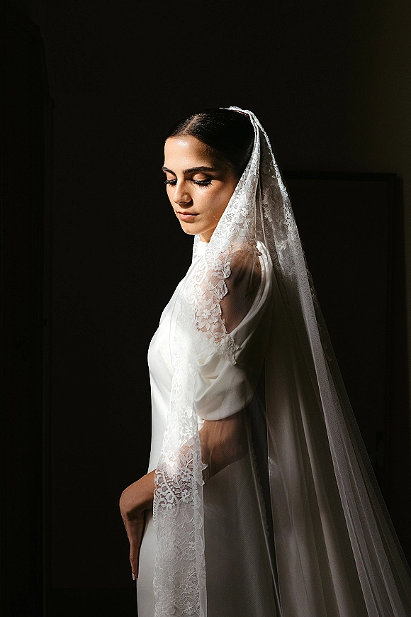 Bridal portrait of a bride in a lace wedding veil and white dress with lace sleeves, looking down in window light against a dark wall