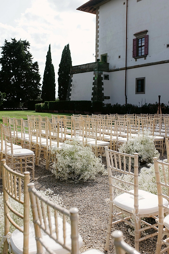 Ceremony setup with chiavari chairs and white cushions lining a gravel aisle with baby's breath clusters outside a white villa lawn