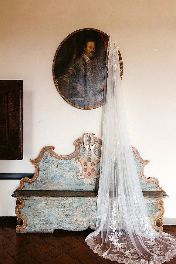 Wedding veil with lace cathedral veil trim draped over an antique painted bench, paired with pointed bridal shoes beneath an oval portrait