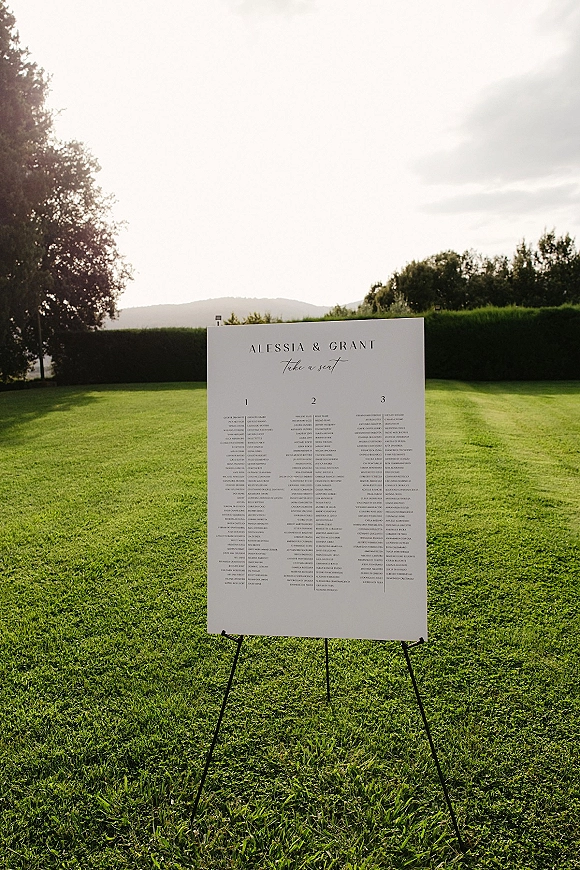 Wedding seating chart sign on an easel, a minimalist black-and-white board displayed on a grass lawn with hedges and hills beyond