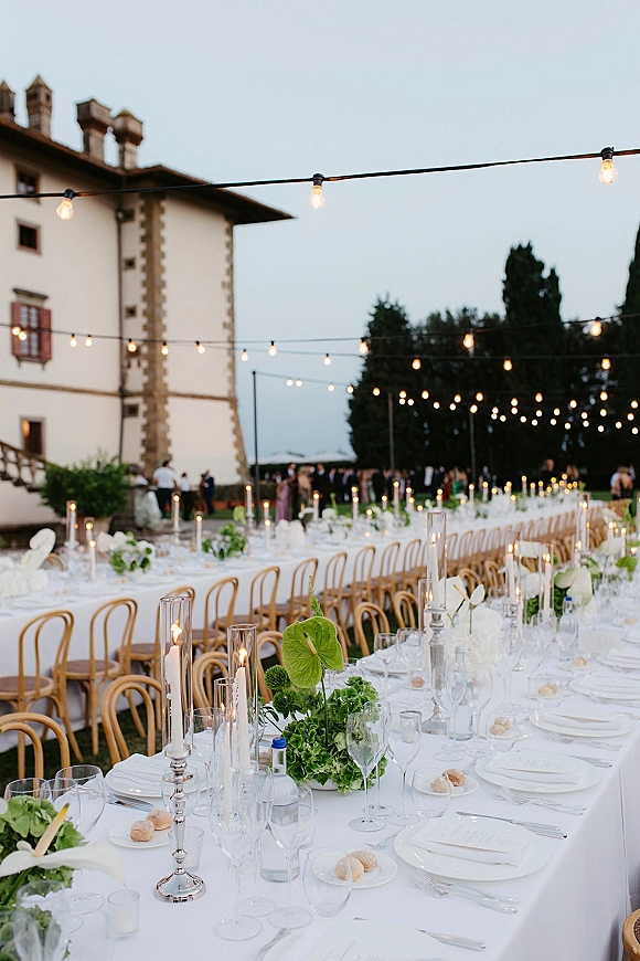 Reception tablescape with long banquet table wedding details, green floral centerpieces and taper candles under string lights on a villa lawn at dusk