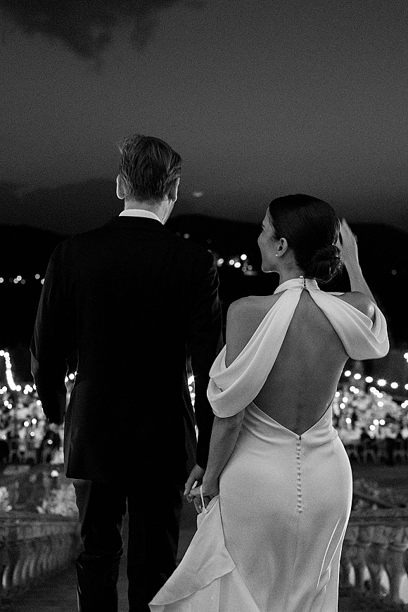 Couple portrait in a black and white wedding photo, bride and groom from behind under bistro string lights at an outdoor reception at night