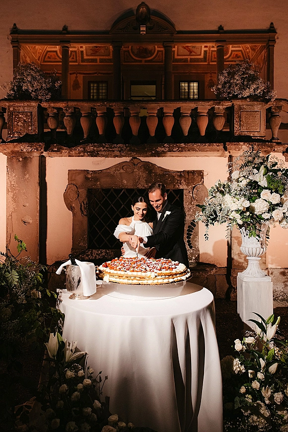 Wedding cake cutting as bride and groom slice a berry-topped cake on a white tablecloth, with white rose floral accents and stone wall backdrop