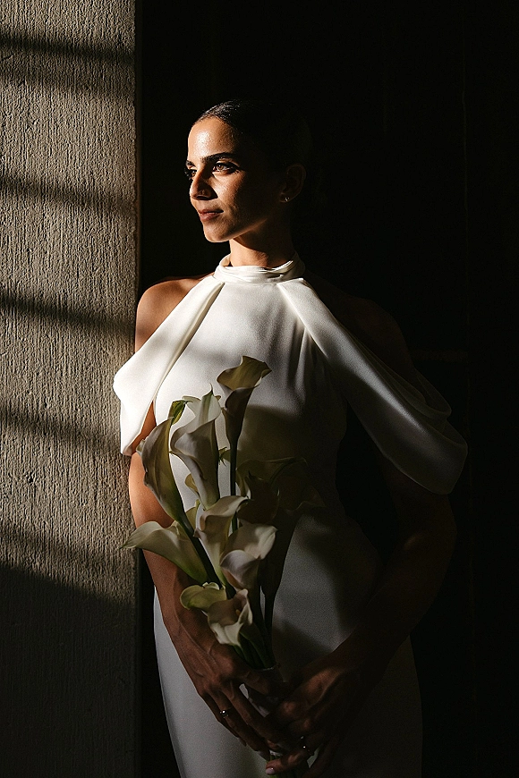 Bridal portrait of a bride holding bouquet of white calla lilies, wearing a high-neck white dress in window light against a shadowy wall.