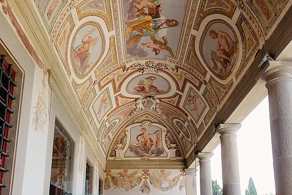 Ornate fresco ceiling with painted ceiling fresco panels and decorative plaster molding above arched corridor with stone columns and light beyond