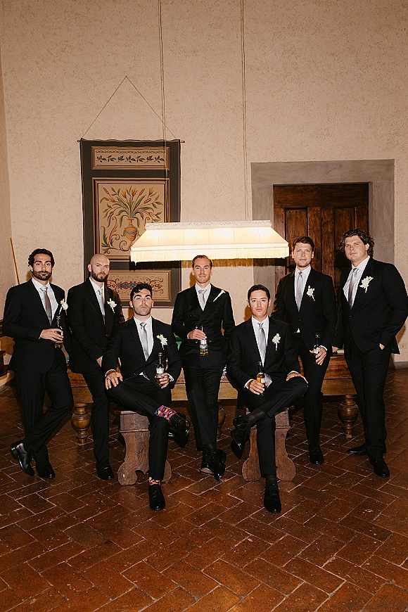 Groomsmen portrait of a men wedding party in black suits with white ties and boutonnieres, holding beer bottles on a wooden bench indoors