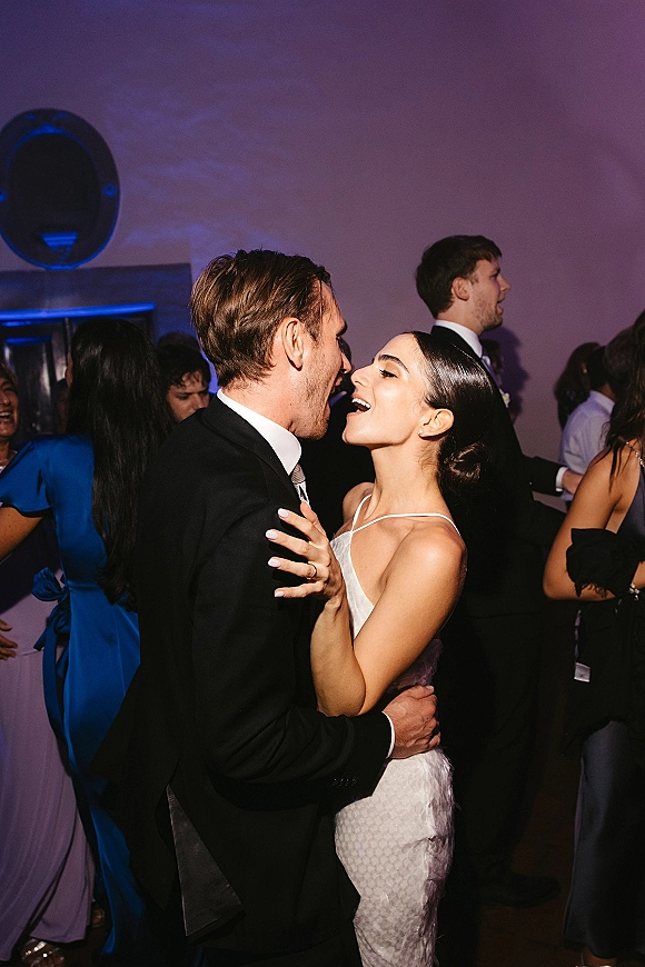 Wedding dance moment as bride and groom dancing in close embrace, smiling on a reception dance floor with purple uplighting and guests behind