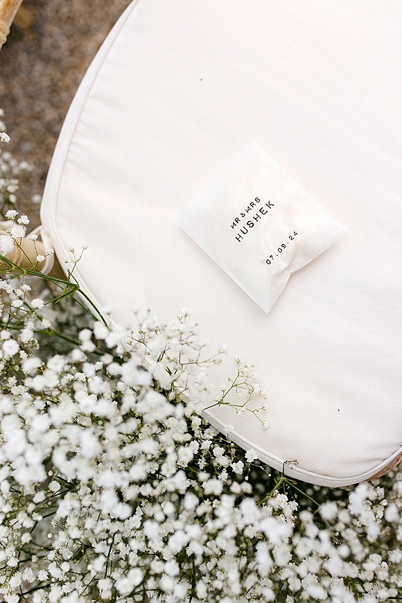 Wedding favor bag with custom wedding favors text, printed names and date, tied with baby’s breath on a white chair cushion above gravel