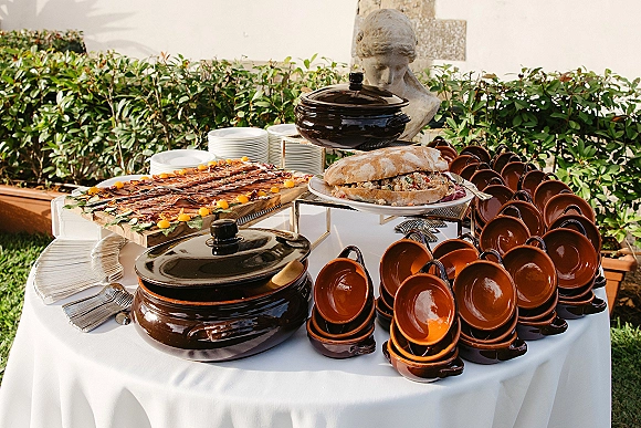 Wedding buffet table with wedding catering display on a white tablecloth, bread loaf, sliced meats, covered bowls, and plates by a garden hedge