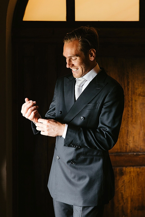 Groom portrait in window light wearing a double breasted wedding suit with white tie, adjusting cufflinks by a dark wood doorway indoors