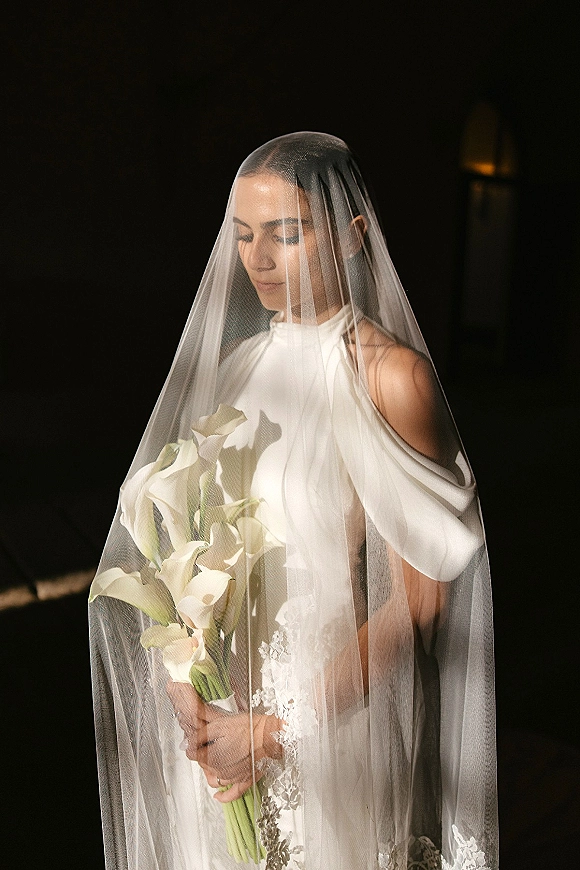 Bridal portrait of a bride with veil holding a white calla lily bouquet, softly lit by a spotlight in a dark interior setting