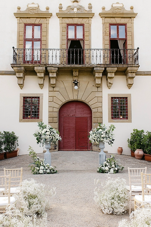 Ceremony altar decor with white floral urns and greenery accents framing a stone doorway with red double doors in a gravel courtyard