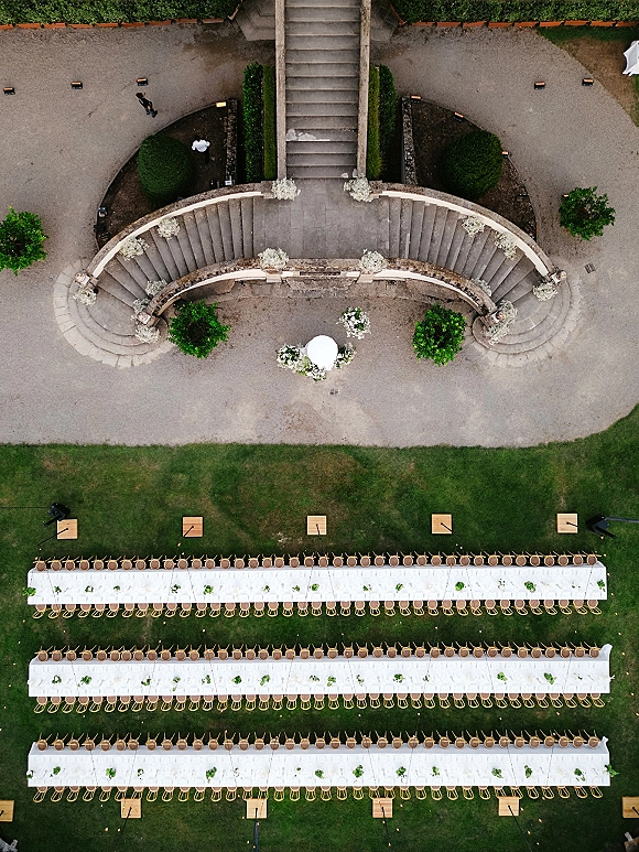 Outdoor reception setup with long banquet table wedding rows in white tablecloths, wooden chairs and string lights on a grassy lawn by stone steps