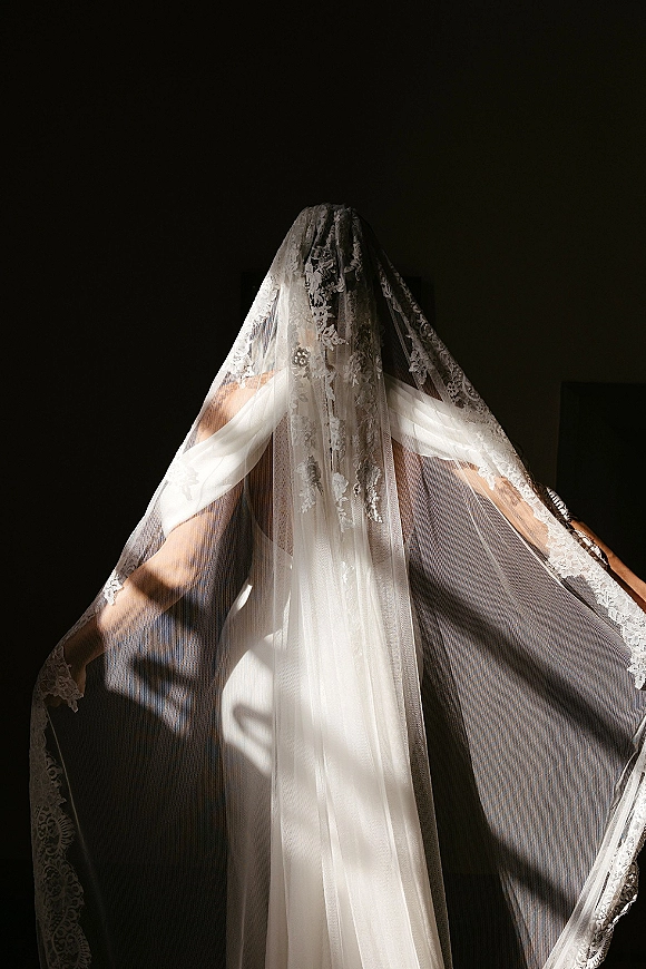 Bridal veil portrait with cathedral length veil and lace mantilla detail, sheer tulle draped in window light against a dark wall