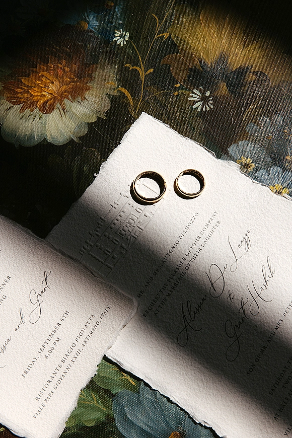Wedding rings, gold wedding bands resting on a deckled edge letterpress invitation with calligraphy on dark floral fabric background