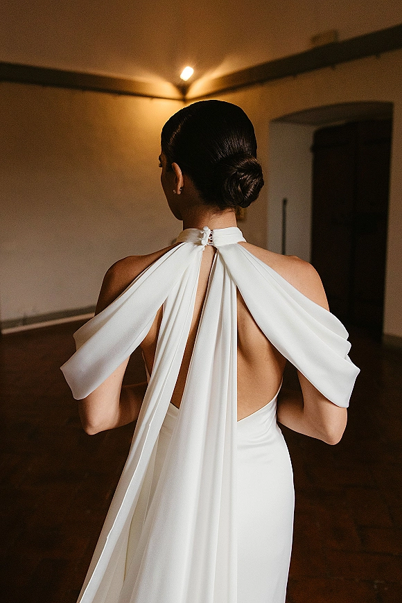 Wedding dress back view with an open back wedding dress, halter neckline and scarf tie, bride in a low bun in an indoor hallway with sconces