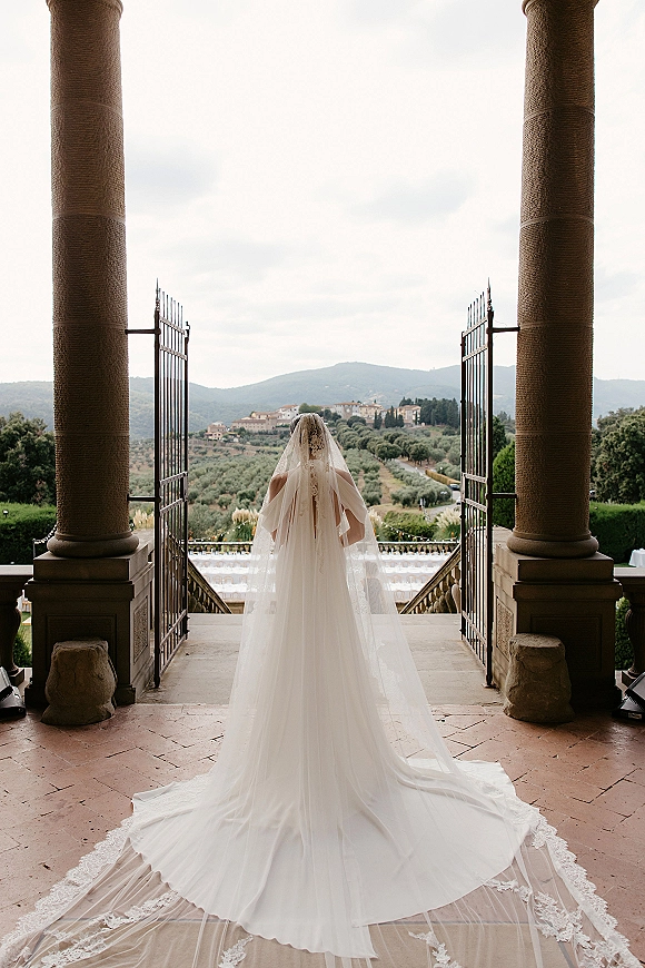 Bridal portrait of a back view bride with cathedral veil and lace train at open iron gates, stone columns framing countryside hills beyond