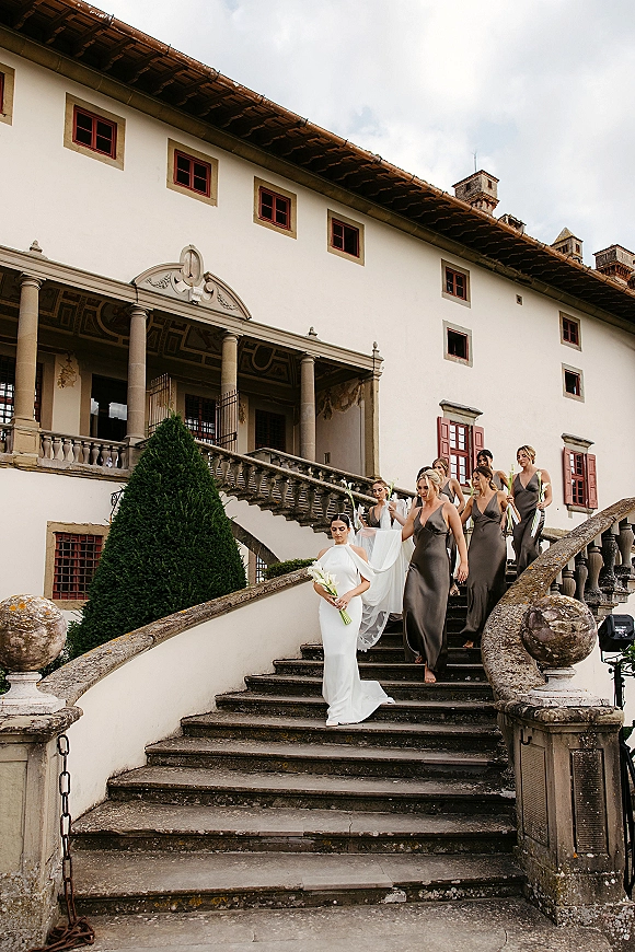 Wedding party entrance as the bride walks down stairs with bridesmaids, holding calla lilies on a stone staircase at a historic villa facade