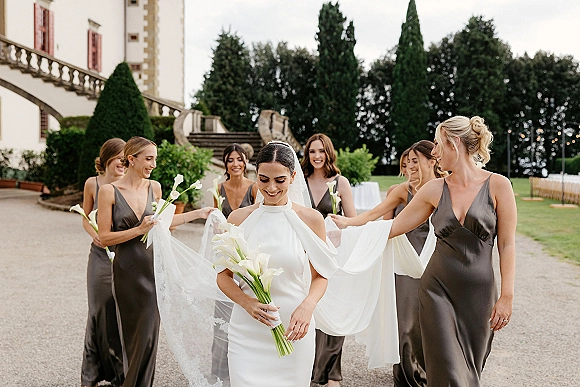 Bride with bridesmaids walking down a stone staircase, bridesmaids holding her long veil and calla lily bouquets outside a mansion