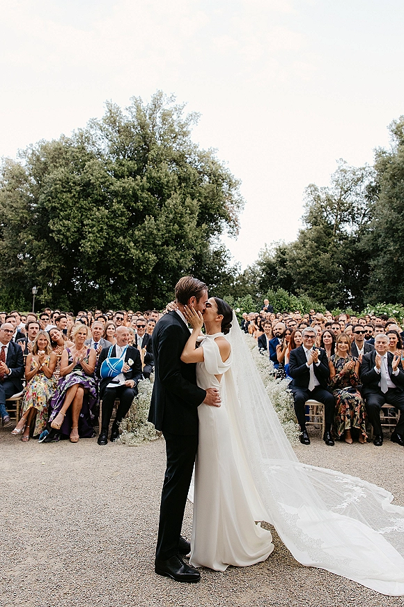 Wedding kiss as bride in off-shoulder dress and cathedral veil train embraces groom in black suit on a gravel garden aisle with guests seated
