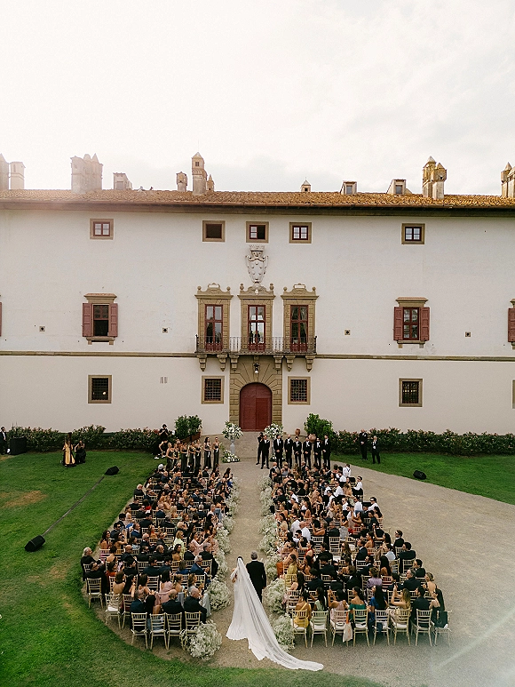 Wedding ceremony in an outdoor courtyard ceremony as the bride walks down a gravel aisle with a long cathedral veil and white flowers, guests seated by a historic facade
